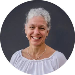 Porträt von lächelnder Ingrid Wimmer mit grauem Lockenhaar, weißer Bluse und Perlenkette vor dunklem Hintergrund.