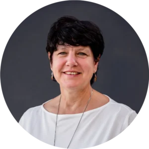 Porträt von Maria Wimmer mit kurzem dunklem Haar, weißer Bluse und Kette vor dunklem Hintergrund, freundlich lächelnd.