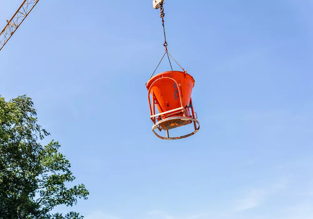Eine orangefarbene Betonkübel-Hebevorrichtung hängt an einem Kran vor blauem Himmel, links sind Baumkronen zu sehen.