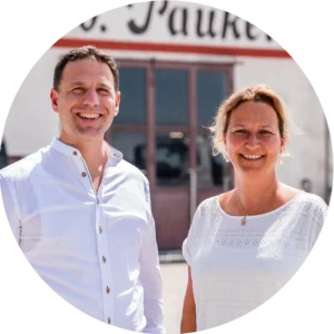 Sebastian und Petra Pauker lächeln in die Kamera, beide in weißer Kleidung, vor einem Gebäude mit der Aufschrift ‚Pauker‘.