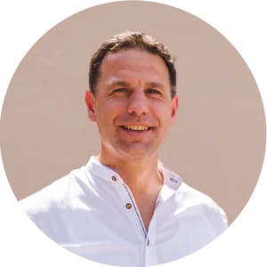 Lächelnder Sebastian Pauker in weißem Hemd vor beigefarbener Wand, frontal aufgenommen.