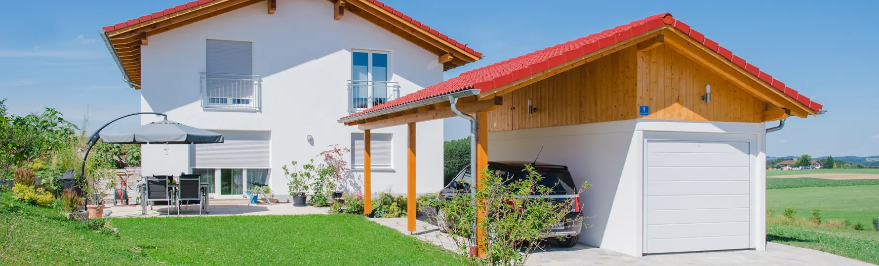 Zweistöckiges Einfamilienhaus mit weißer Fassade und rotem Satteldach, angrenzend freistehende Garage mit rotem Dach, gepflegter Garten mit Terrasse und Gartenmöbeln.
