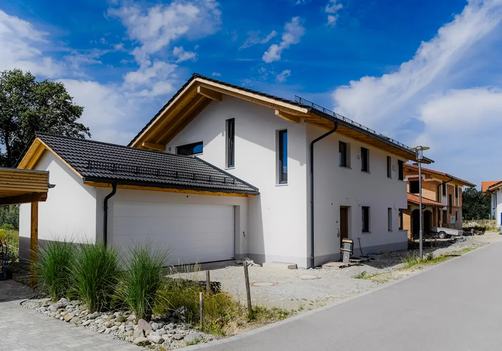 Modernes Einfamilienhaus mit Garage und dunklem Ziegeldach, Kiesfläche im Vorgarten, Neubaugebiet im Hintergrund.