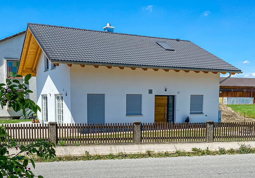 Modernes Einfamilienhaus mit weißer Fassade, grauem Dach und Holztür, umgeben von einem braunen Holzzaun.