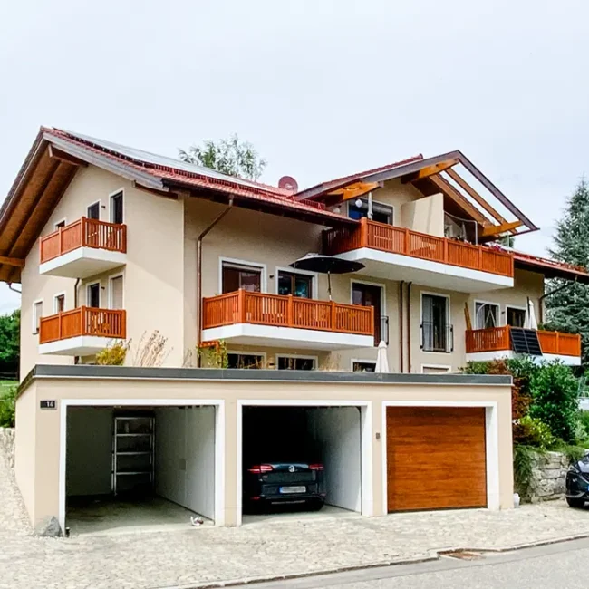 Mehrfamilienhaus mit Holz-Balkonen, Solarpanel und drei Garagen; davor geparkte Autos auf gepflasterter Fläche.