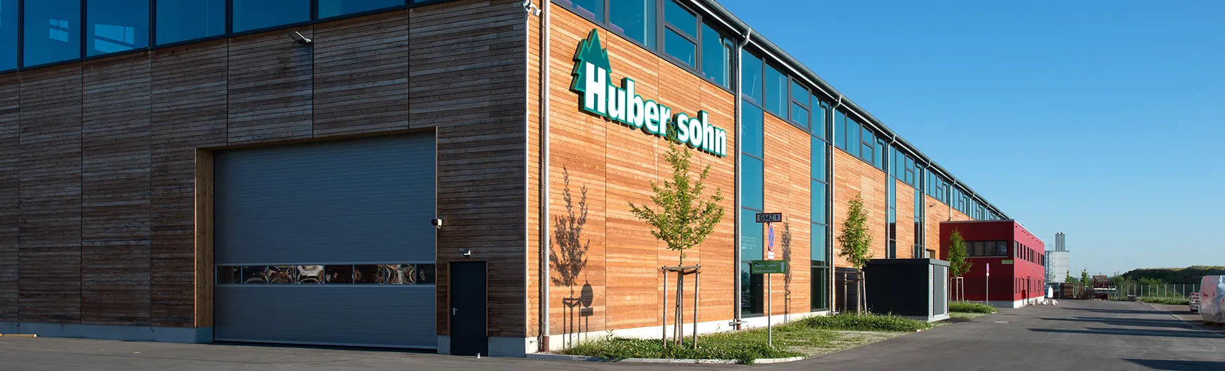 Moderne Industriehalle mit Holzfassade und dem Logo ‚Huber & Sohn‘, daneben rotes Bürogebäude und breite Zufahrt.