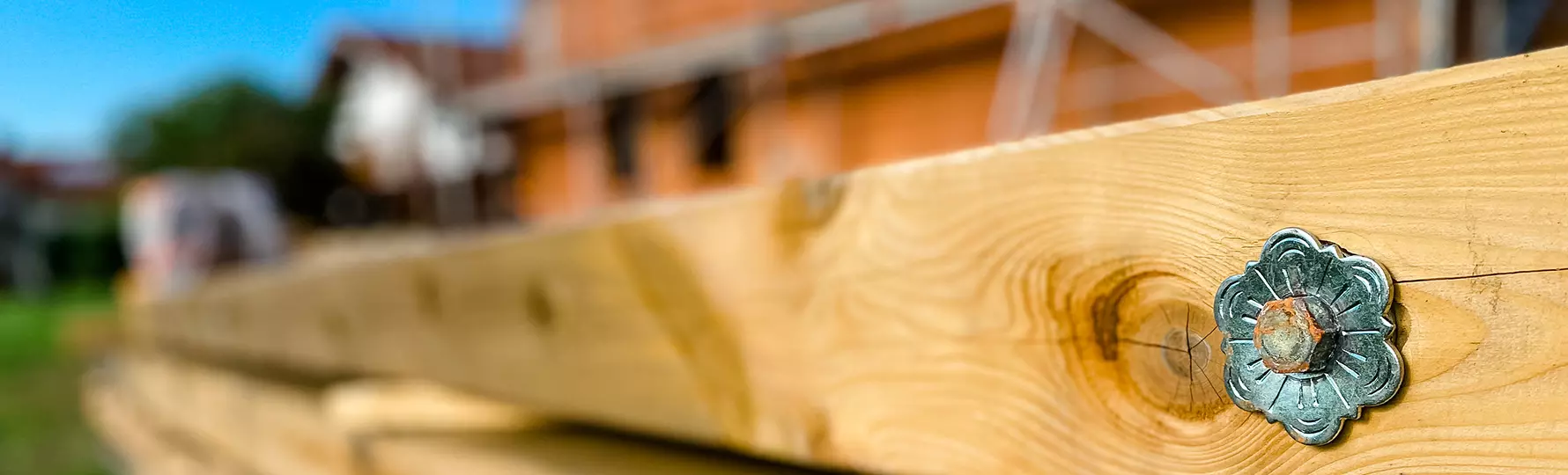 Nahaufnahme eines Holzbalkens mit dekorativer Metallschraube, unscharfer Rohbau im Hintergrund.