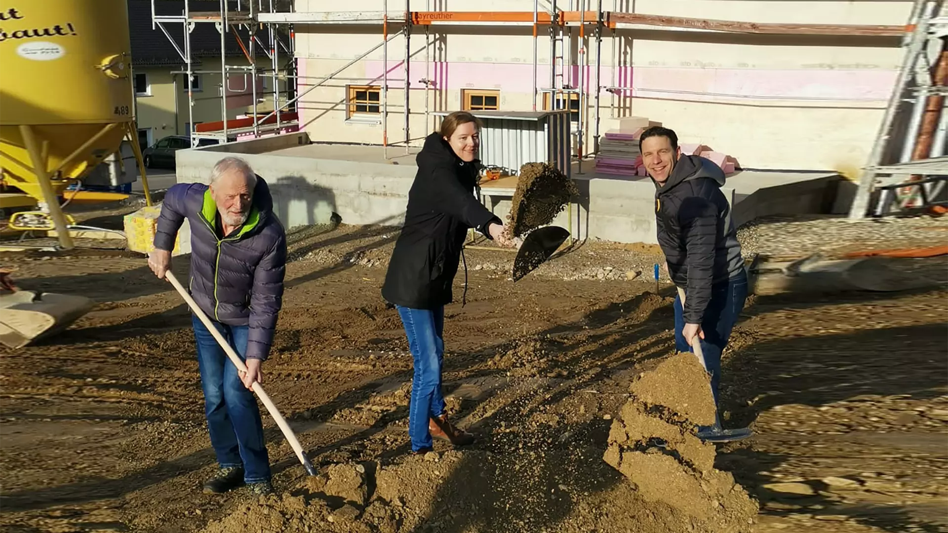 Drei Personen mit Spaten beim symbolischen Spatenstich auf einer Baustelle vor einem Rohbau und Baugeräten.