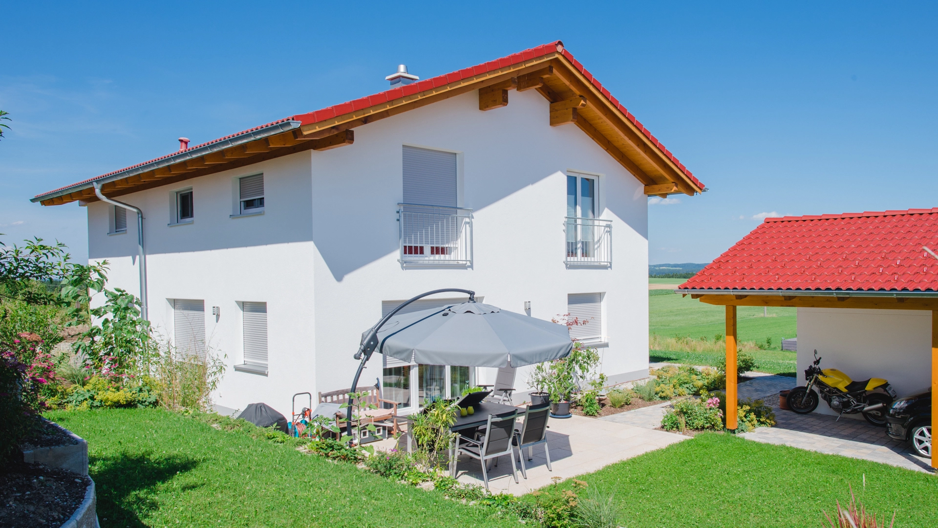 Modernes weißes Einfamilienhaus mit rotem Satteldach, Terrasse mit Sitzgruppe und angrenzendem Carport.