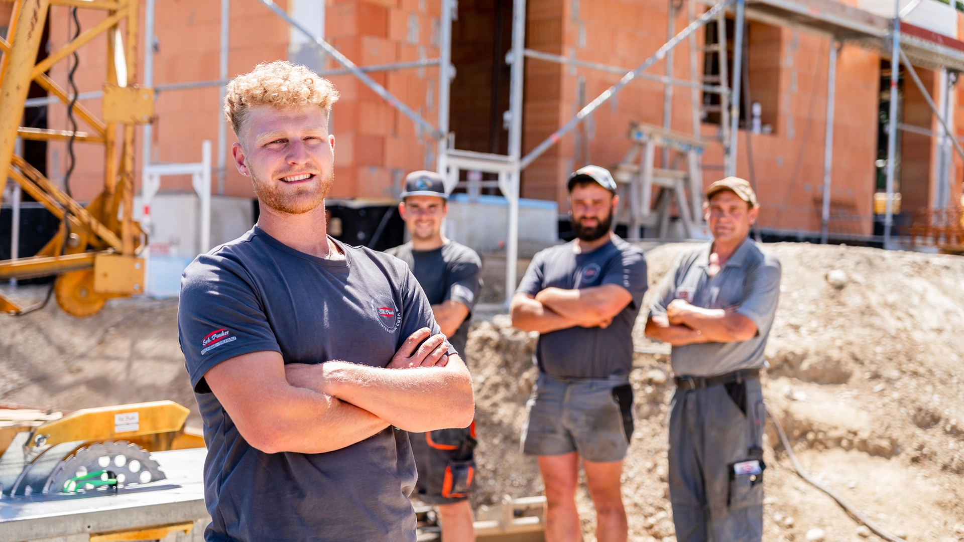 Vier Bauarbeiter mit verschränkten Armen vor einer Baustelle mit unverputztem Ziegelbau und Baugerüst.