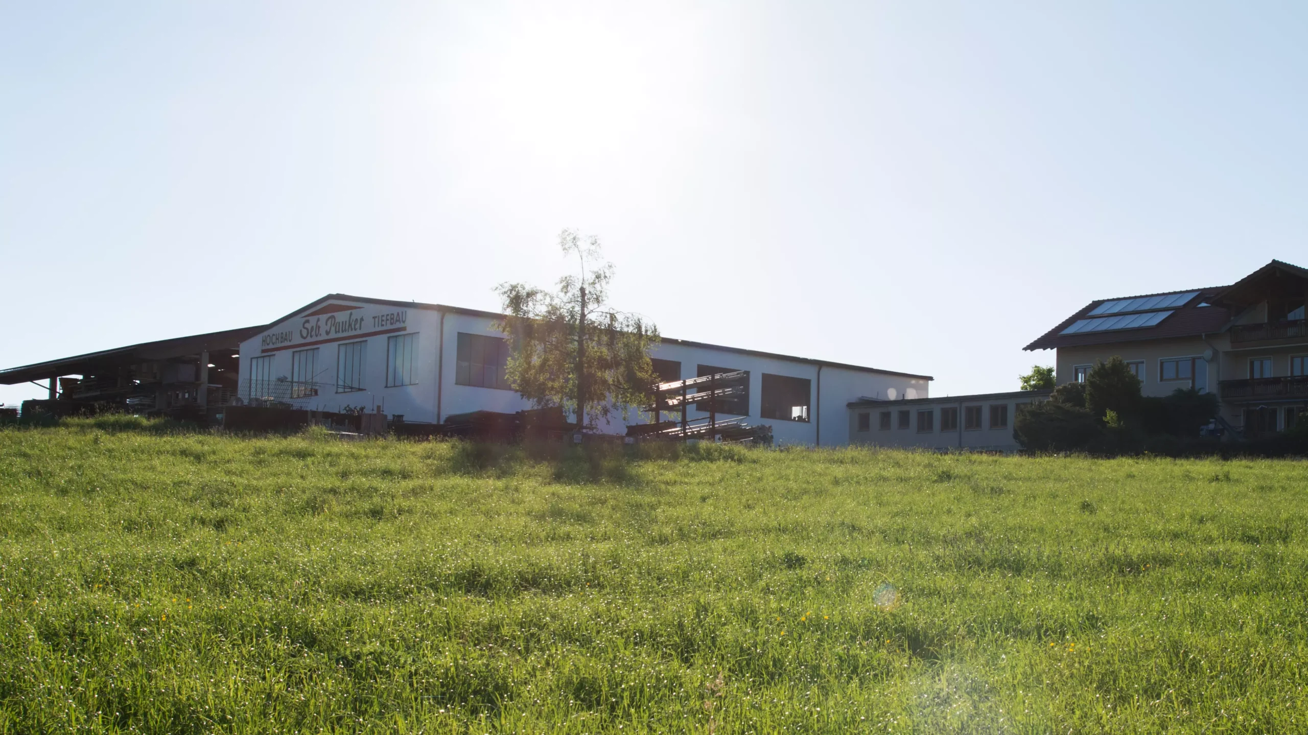Gewerbegebäude mit Aufschrift „Hochbau Tiefbau Seb. Pauker“ auf Wiese, im Gegenlicht neben Wohnhaus mit Solaranlage.
