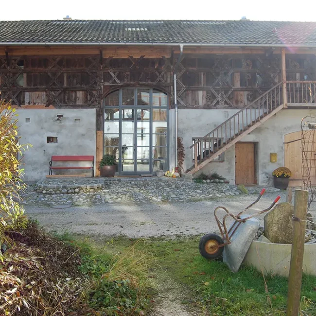 Vorderansicht eines renovierten Bauernhauses mit Steinmauer, Holzfassade und Treppe, davor eine rote Bank, ein Pflanztopf und eine Schubkarre neben einem alten Brunnen.