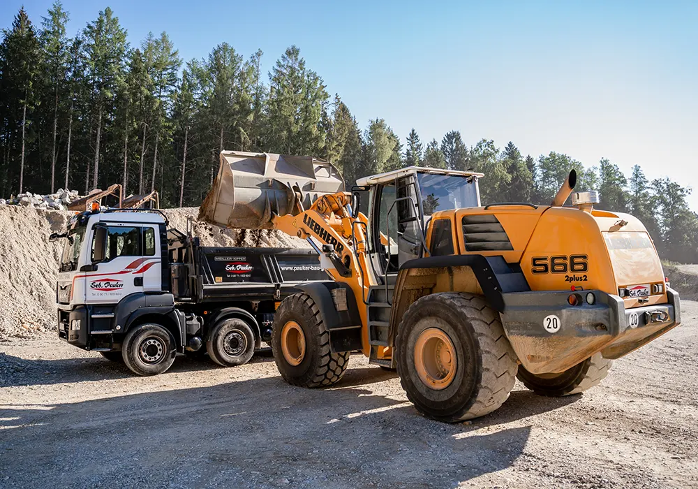 Gelber Radlader lädt Erdmaterial in Lkw mit Aufschrift „Seb. Pauker“ auf Baustelle vor Waldrand.