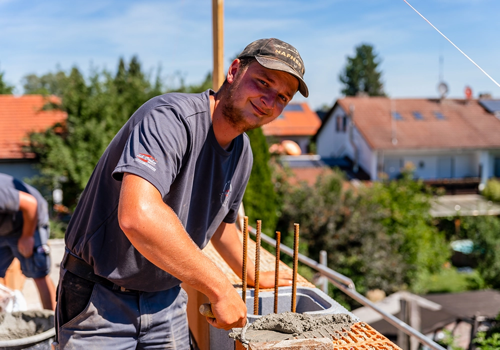 Bauarbeiter trägt Mörtel auf eine Mauer mit Armierungseisen auf und lächelt in die Kamera.