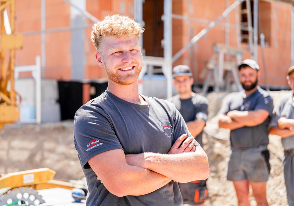 Junger Bauarbeiter mit verschränkten Armen lächelt vor Kollegen auf einer Baustelle in die Kamera.