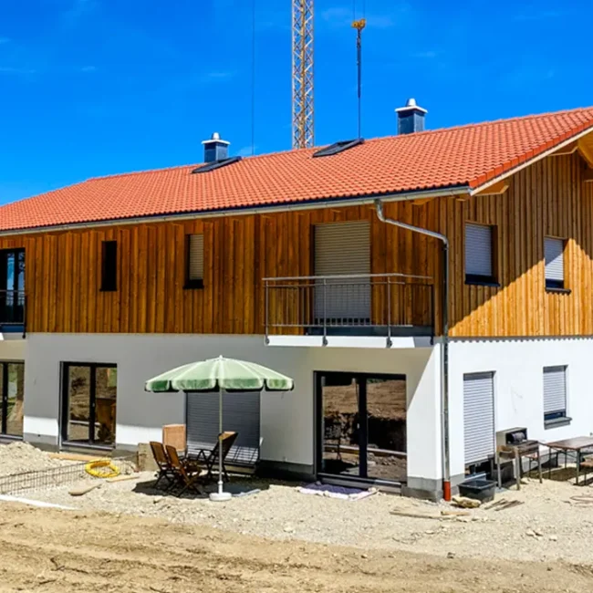 Zweigeschossiges Wohnhaus im Bau mit rotem Ziegeldach, Holzfassade im Obergeschoss, weißem Putz im Erdgeschoss und möblierter Terrasse mit Sonnenschirm.