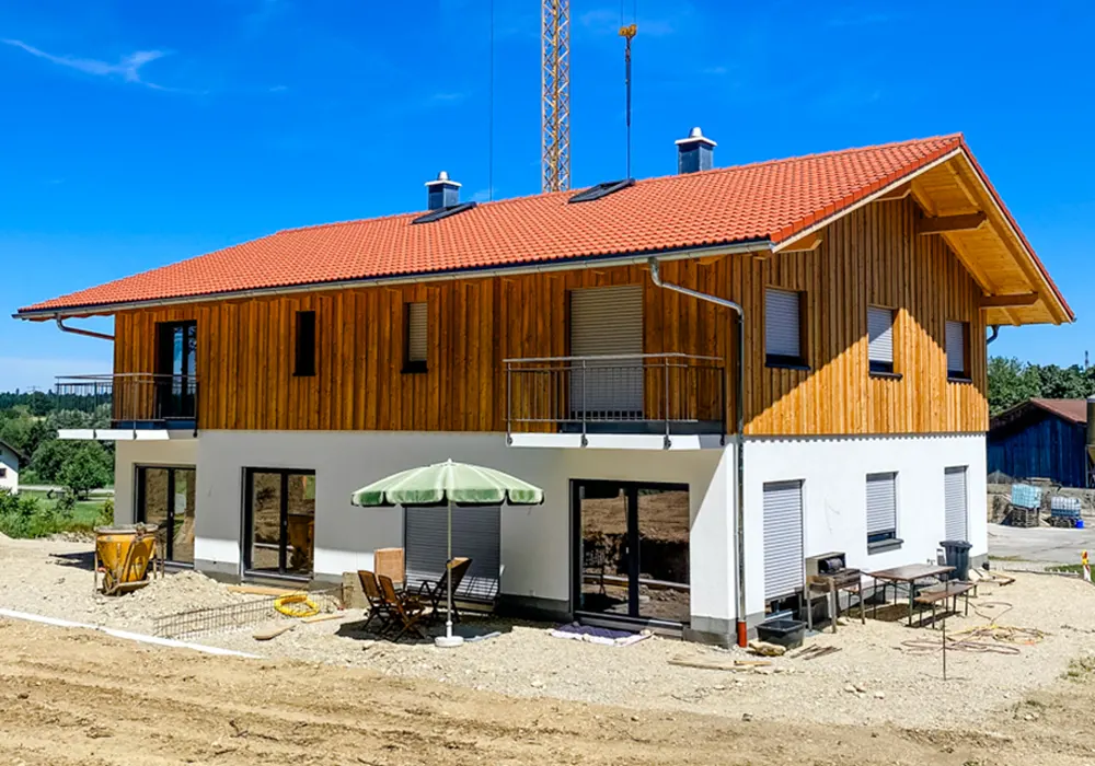 Zweigeschossiges Wohnhaus im Bau mit rotem Ziegeldach, Holzfassade im Obergeschoss, weißem Putz im Erdgeschoss und möblierter Terrasse mit Sonnenschirm.