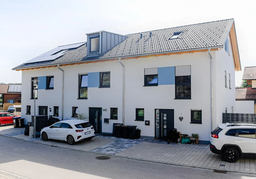 Modernes weißes Doppelhaus mit grauem Dach, blauen Fassadenelementen, Solarpanels und parkenden Autos vor dem Haus.