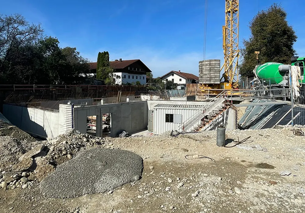 Baustelle mit Betonkeller, Baucontainer, Kran, Zementsilo und grünem Betonmischer vor Wohnhäusern im Hintergrund.
