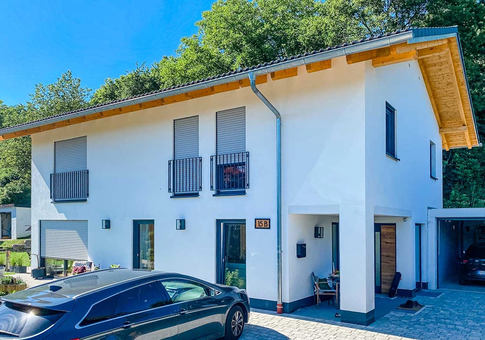 Modernes weißes Wohnhaus mit Holzdetails, Rollläden und Carport, davor zwei parkende Autos.