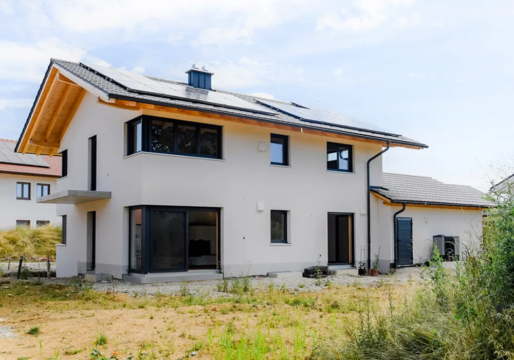 Modernes zweigeschossiges Einfamilienhaus mit heller Putzfassade, großen Fenstern, Solarpanels auf dem Dach und naturbelassenem Garten im Vordergrund.