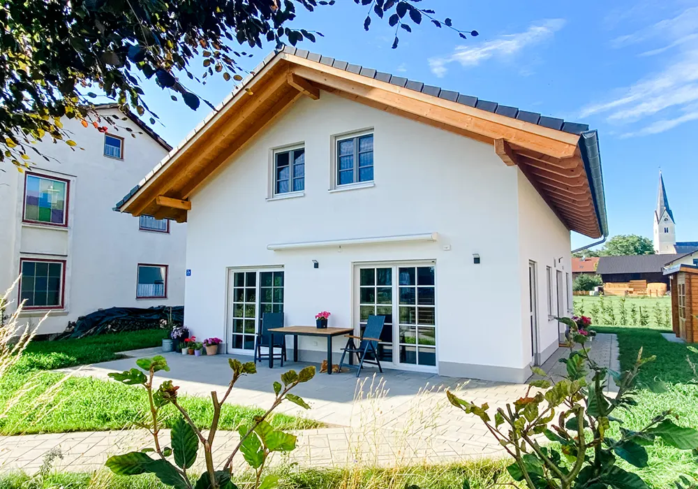 Modernes Einfamilienhaus mit weißer Fassade, Holzdach, Terrasse und Garten, im Hintergrund ein Kirchturm.