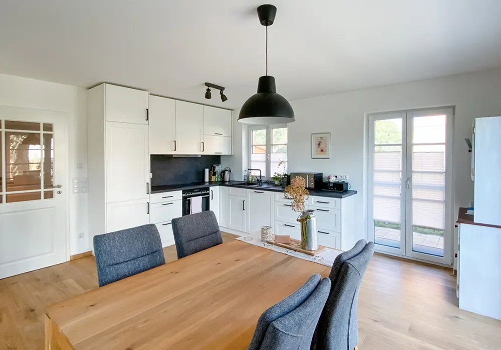 Helle, moderne Wohnküche mit weißen Einbauschränken, schwarzer Arbeitsplatte, Holz-Esstisch und großen Fenstern.