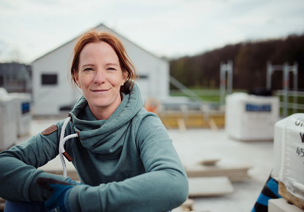 Frau mit roten Haaren und grünem Kapuzenpullover lächelt in die Kamera auf einer Baustelle.