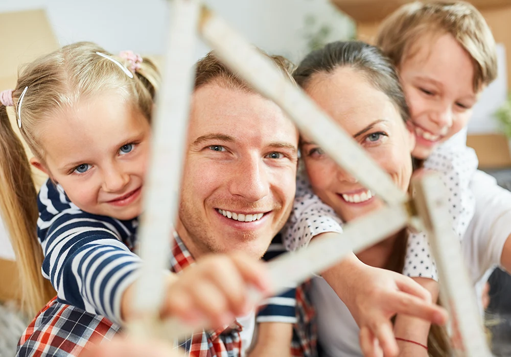 Lächelnde Familie mit zwei Kindern blickt durch einen aufgeklappten Zollstock in Form eines Hauses.