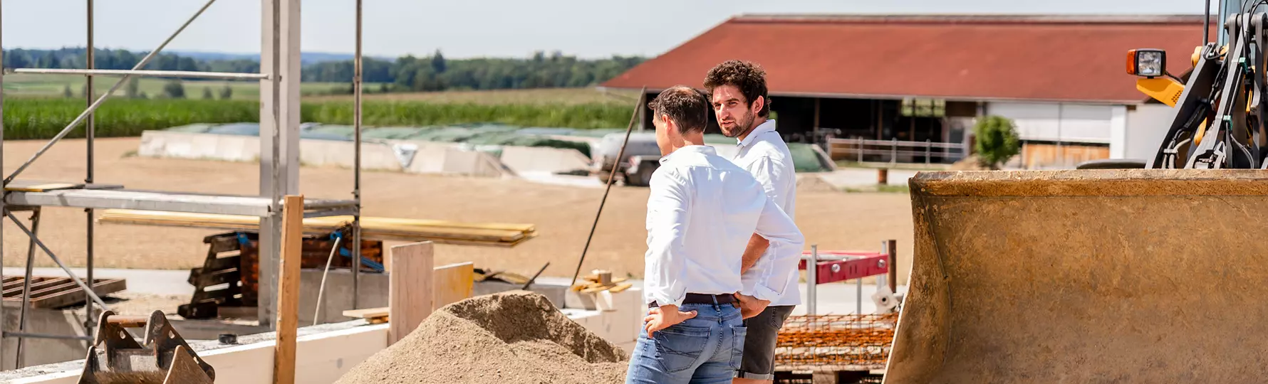 Zwei Männer in weißem Hemd stehen auf Baustelle neben Baggerlöffel und Sandhaufen im Gespräch.