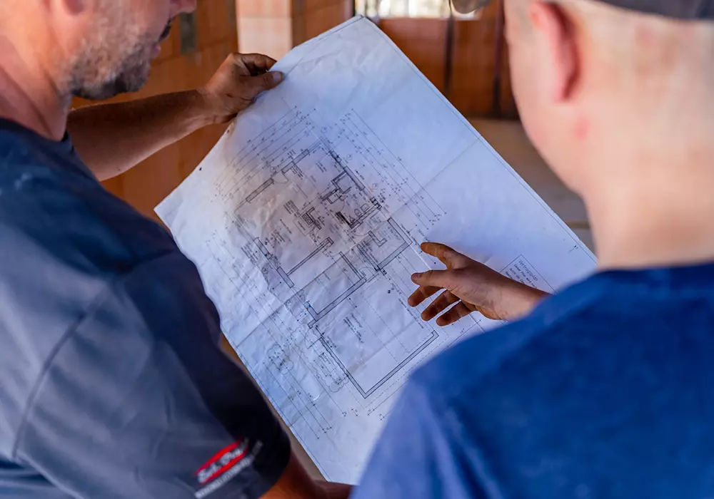 Zwei Männer betrachten einen Bauplan; einer zeigt mit dem Finger auf eine Stelle auf der Zeichnung.