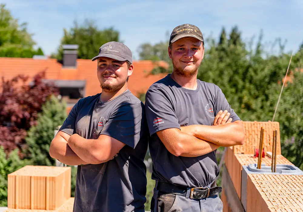 Zwei junge Bauarbeiter mit verschränkten Armen posieren auf einer Baustelle vor Mauer aus Ziegelsteinen.