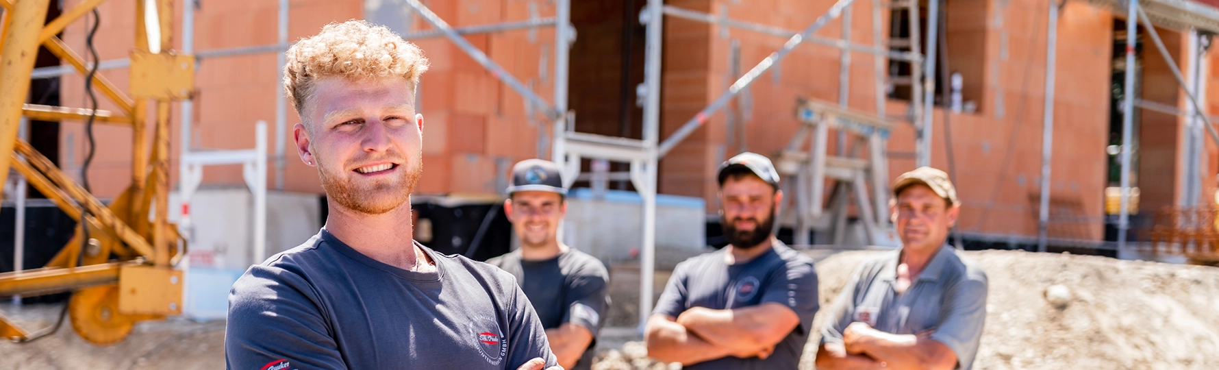 Vier Bauarbeiter mit verschränkten Armen vor einer Baustelle mit unverputztem Ziegelbau und Baugerüst.