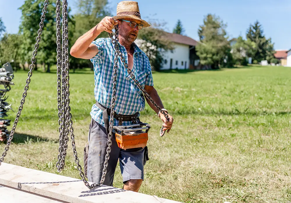 Mann mit Strohhut befestigt Kette an Last auf Wiese, trägt Funksteuerung für Kran um die Hüfte.
