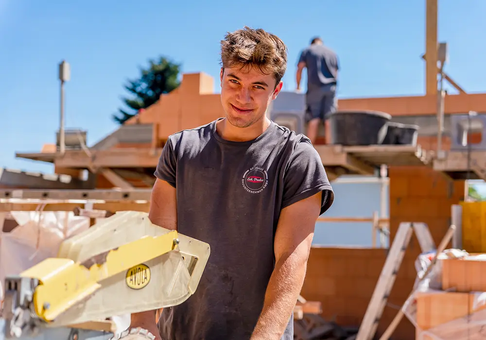 Ein junger Bauarbeiter steht lächelnd vor einer Baustelle, vor ihm eine Kappsäge; im Hintergrund arbeitet eine weitere Person auf einem Gerüst.