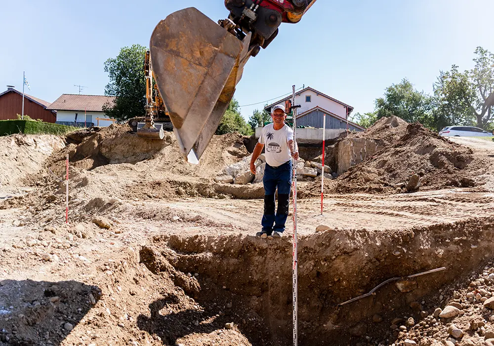 Bauarbeiter misst Tiefe einer Baugrube mit Messlatte, im Vordergrund Baggerlöffel, im Hintergrund Erdhügel.