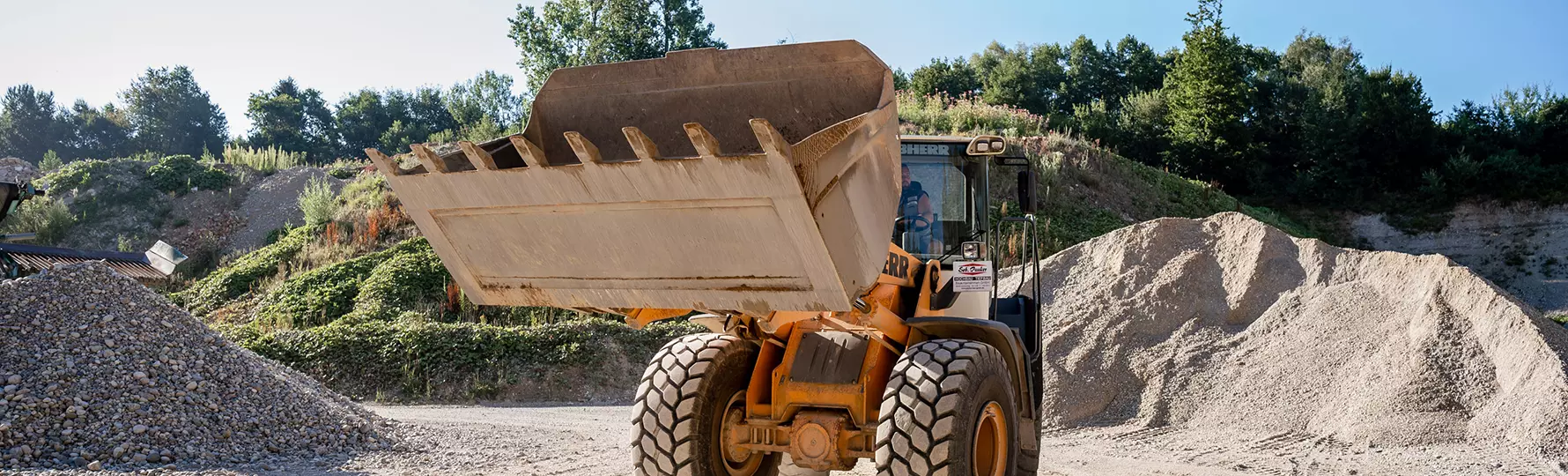 pauker-bau-gmbh-kiesgrube-header Radlader mit erhobener Schaufel auf einem Kiesplatz, im Hintergrund Kieshaufen und bewaldete Böschung unter blauem Himmel.