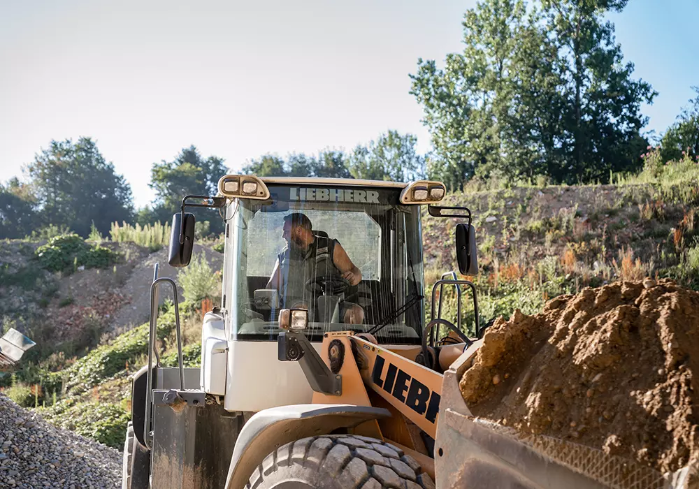 pauker-bau-gmbh-kiesgrube-vorteile-03 Radlader der Marke Liebherr mit beladener Schaufel, ein Mann sitzt in der Fahrerkabine und schaut nach links; im Hintergrund ein Hang mit Vegetation und ein Kieshaufen.
