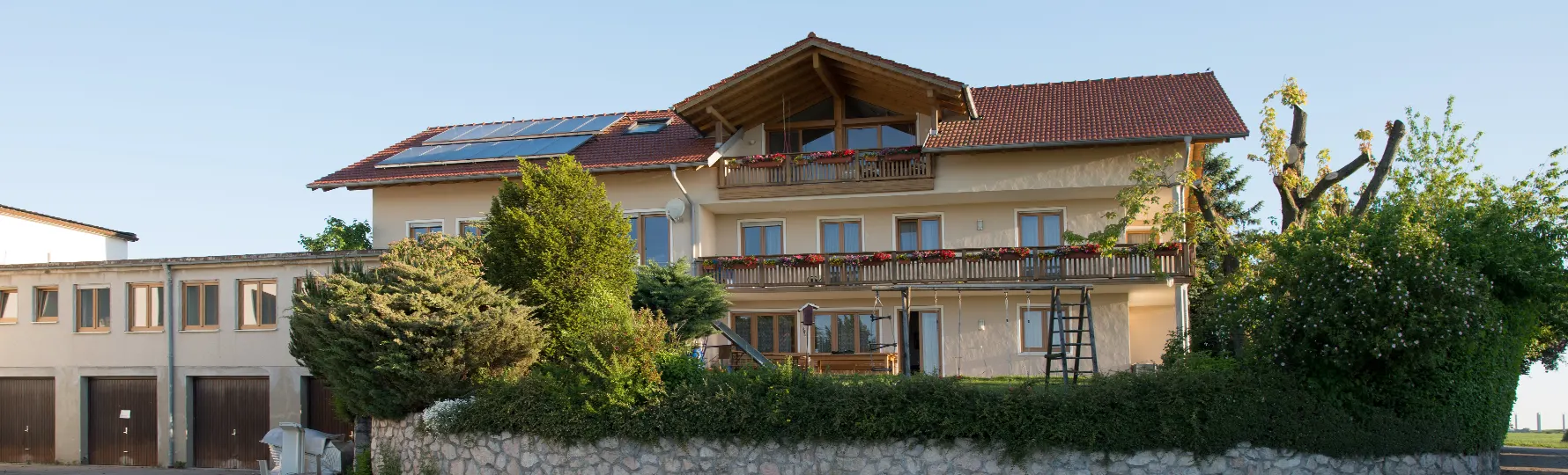 pauker-bau-gmbh-kontakt-header Zweistöckiges Wohnhaus mit rotem Satteldach, Balkonen mit Blumenkästen und Solarpaneelen auf dem Dach, daneben Garagengebäude, umgeben von Bäumen und Sträuchern.