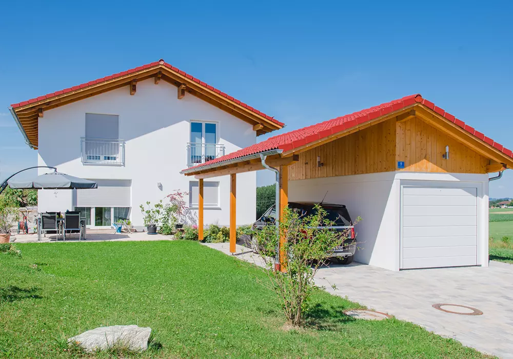 Modernes Einfamilienhaus mit rotem Satteldach, Garten, Terrasse und freistehender Garage mit Carport und geparktem Auto.