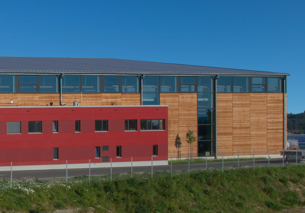 Moderne Halle mit Holzfassade und rotem Bürotrakt, davor Straße und Zaun.
