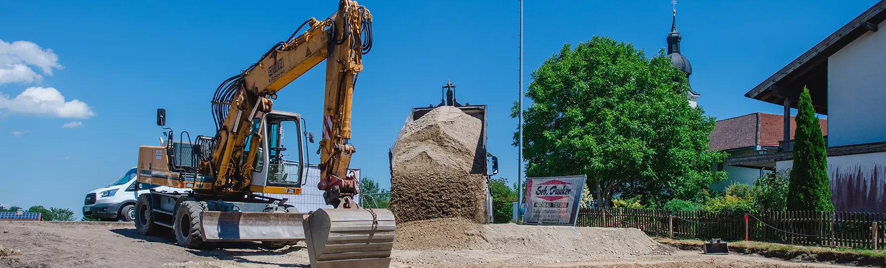 Bagger von Seb. Pauker Hochbau Tiefbau GmbH beim Aushub neben LKW mit Erdhügel, Bauzaun mit Firmenschild im Hintergrund.