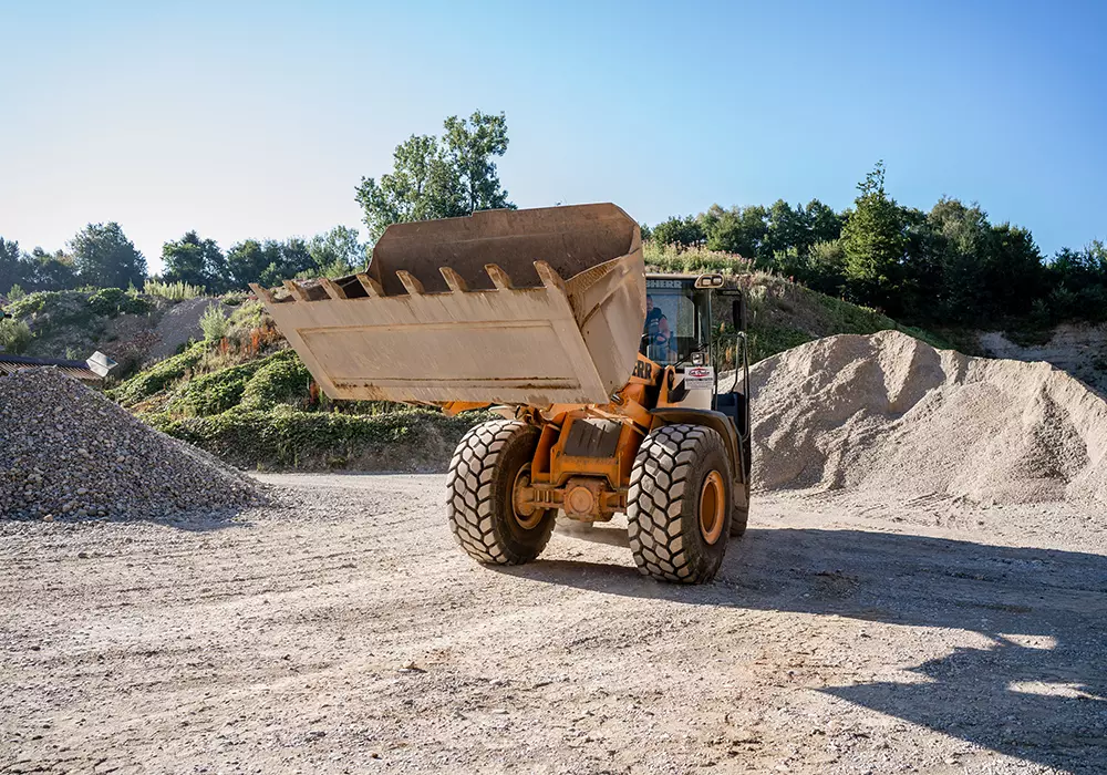 Radlader mit erhobener Schaufel auf einem Kiesplatz, im Hintergrund Kieshaufen und bewaldete Böschung unter blauem Himmel.