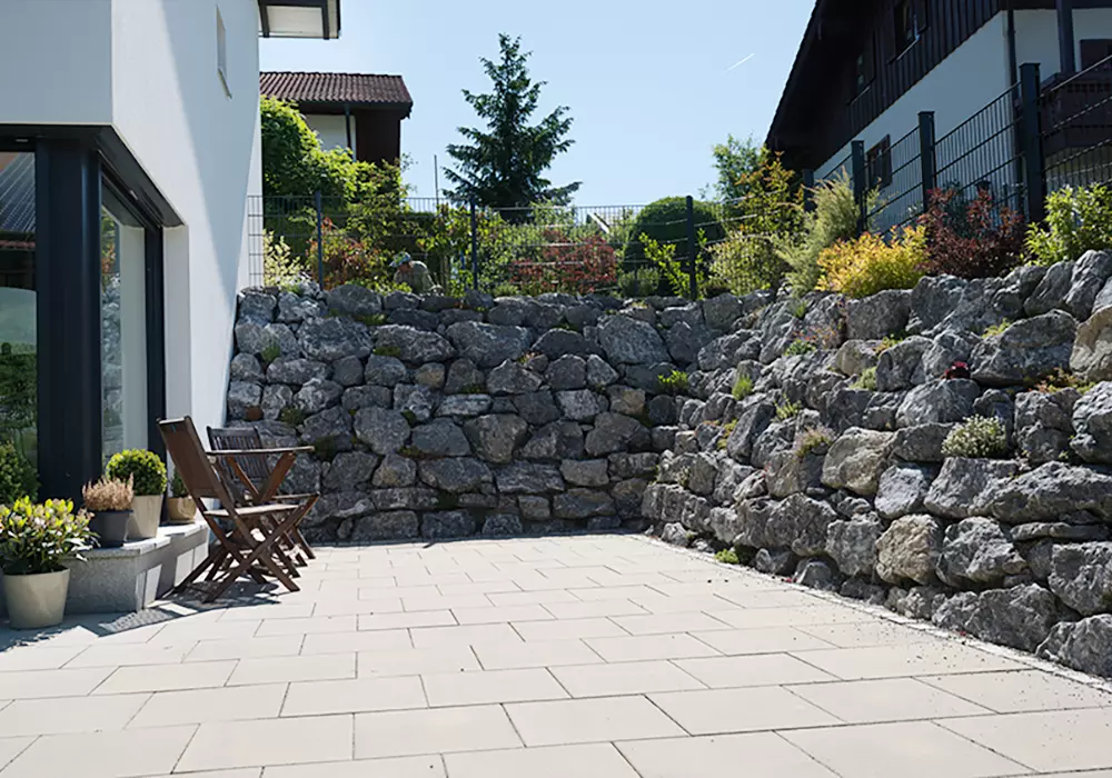 Terrasse mit Holzstühlen vor einem modernen Haus, umgeben von bepflanzten Natursteinmauern als Hangbefestigung.