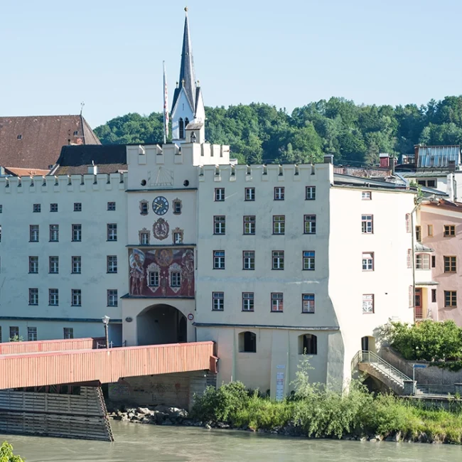Historisches Gebäude mit Uhrturm und bemalter Fassade an Flussufer, davor eine überdachte Holzbrücke.
