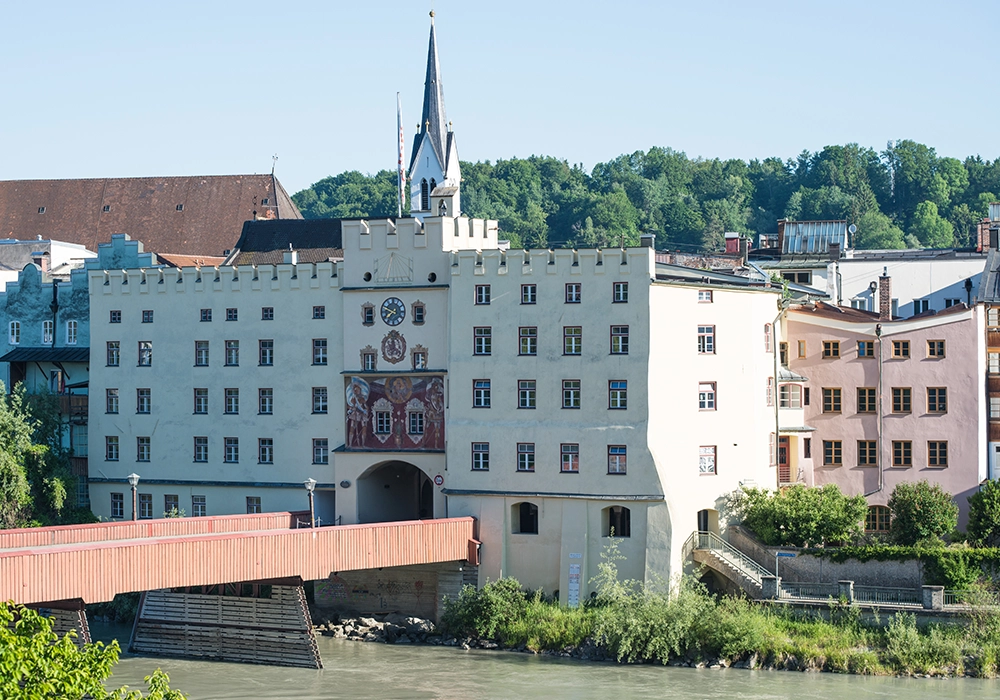 Historisches Gebäude mit Uhrturm und bemalter Fassade an Flussufer, davor eine überdachte Holzbrücke.