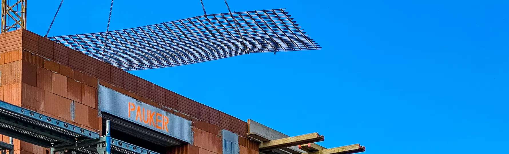 Kran hebt Stahlgitter über Rohbau mit Schriftzug ‚PAUKER‘ auf Ziegelwand.