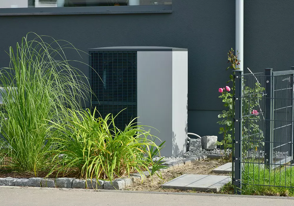 Moderne Wärmepumpe steht außen neben einem Haus, umgeben von Gräsern, Blumen und einem Metallzaun.