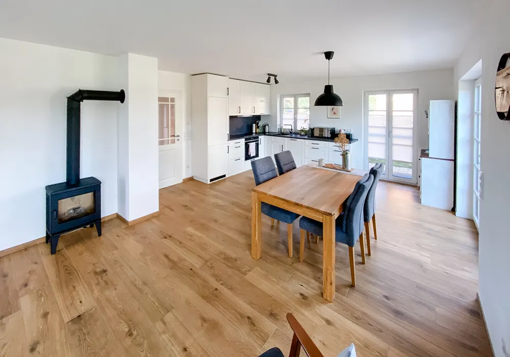 Helles Wohn-Esszimmer mit Holzofen, Esstisch, sechs Stühlen und moderner weißer Einbauküche mit Holzfußboden.
