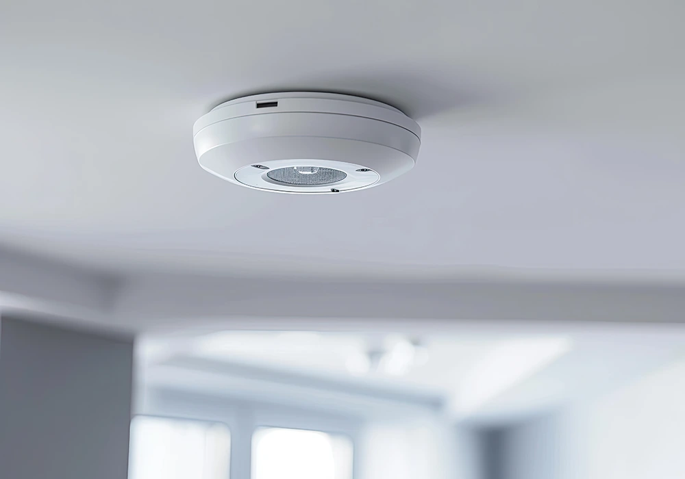 Moderner Rauchmelder oder Sensorgerät an weißer Zimmerdecke in heller Wohnumgebung.